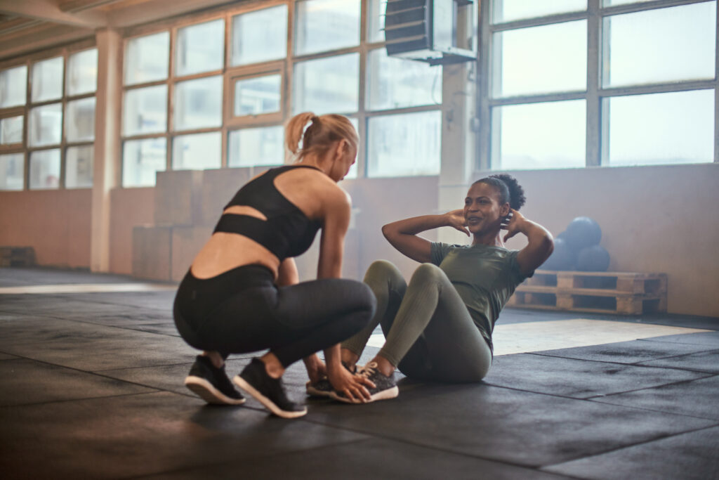 Eine freundlich grinsende Frau macht Situps auf dem Boden eines Fitnessstudios. Eine weitere Frau unterstützt sie dabei.