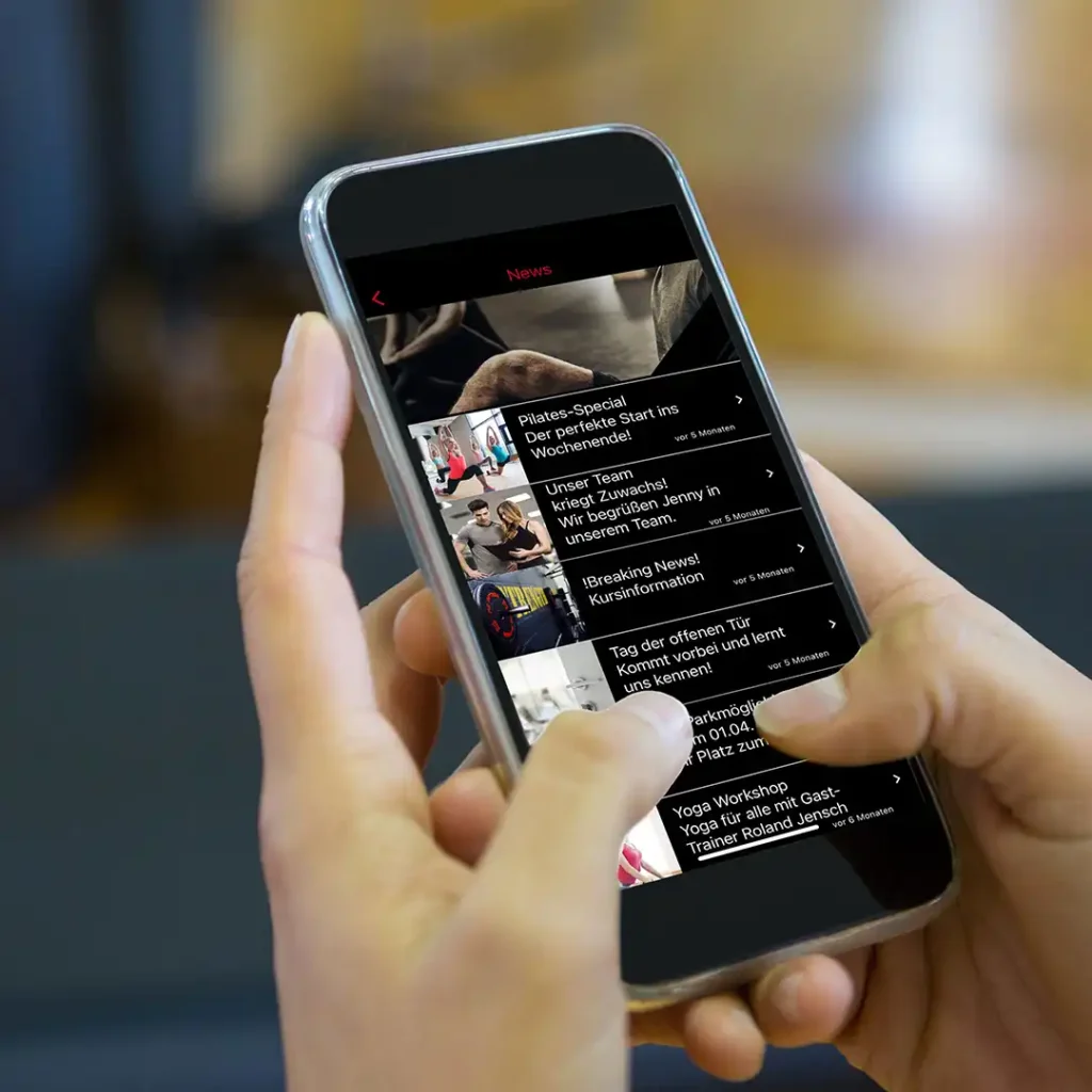 Ein Smartphone mit der Ansicht der CenterCom-App wird in zwei Händen gehalten. Im Hintergrund ist ein Fitnessstudio zu erahnen.