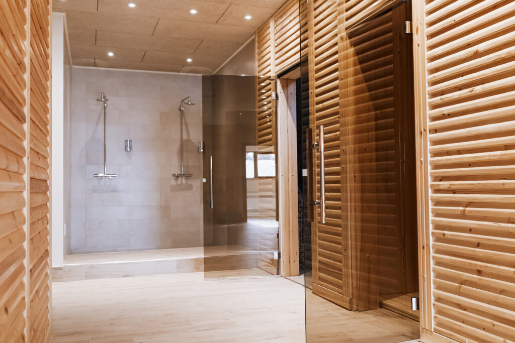 Ein Moderner Spa-Bereich mit zwei offenen Duschen vor heller Fliesenwand, daneben eine Holzsauna mit Glas- und Holztüren, warm beleuchtet.
