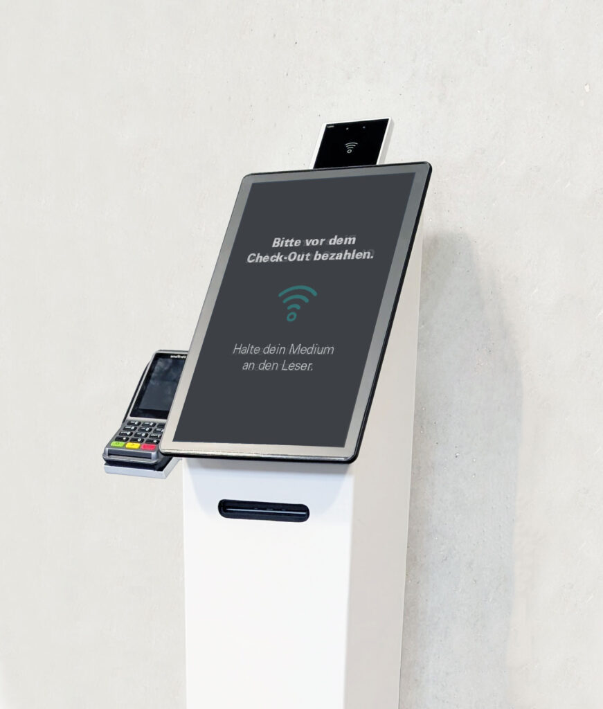 Selbstbedienungs-Terminal mit Touchscreen und Kartenterminal, Anzeige mit NFC-Symbol und Hinweis „Bitte vor dem Check-Out bezahlen. Halte dein Medium an den Leser“, montiert auf weißem Standfuß.
